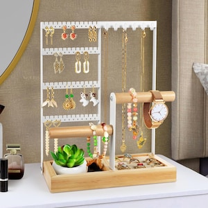 May include: White metal and wood jewelry organizer with multiple earring and necklace displays. Includes a wooden bar for bracelets and a small tray for rings. A gold watch and a small succulent plant are also displayed.