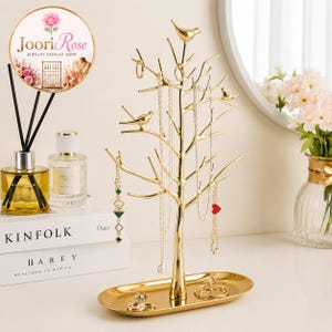 May include: A gold-colored metal jewelry display stand shaped like a tree with branches, featuring small bird figures perched on the limbs. The stand includes an oval base tray for holding rings and small accessories, with several necklaces hanging from the branches.