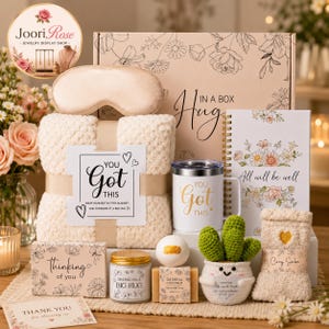 May include: A gift set featuring a beige plush blanket with a You Got This label, a satin sleep mask, a white tumbler with gold text, a spiral notebook with floral art, a candle, a soap bar, a bath bomb, and a crocheted cactus in a pot.