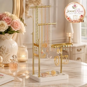 May include: A gold-toned metal jewelry display stand with a white rectangular base, featuring multiple tiers for hanging necklaces, a perforated bar for earrings, and a curved rod for bracelets, holding various necklaces and hoop earrings.