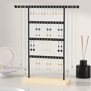 May include: A black metal jewelry display stand with multiple levels, holding various necklaces and earrings. The stand has a wooden base and is adorned with gold, silver, and black jewelry pieces. A small white dish and a black diffuser are on the side.