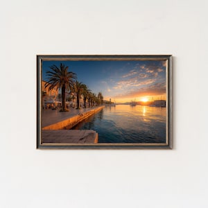 May include: Framed print of a coastal scene at sunset. Palm trees line a stone walkway along the water. Buildings are visible on the left. The sky is a mix of blue, orange, and yellow.