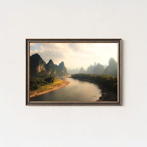 Può includere: Una stampa incorniciata di un paesaggio con un fiume che serpeggia tra montagne verdi sotto un cielo nuvoloso. L'opera d'arte ha una cornice marrone scuro e cattura una scena naturale serena.
