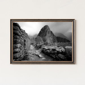 Op de afbeelding: Zwart-wit ingelijste prent van Machu Picchu, met stenen muren, een pad en een berg op de achtergrond. De prent heeft een donkere, sierlijke lijst, die een klassieke touch geeft aan het historische landschap.