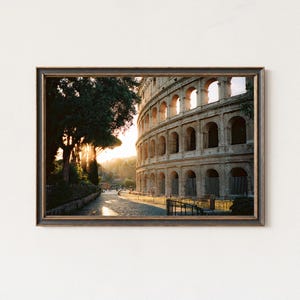 Puede incluir: Impresión enmarcada del Coliseo en Roma, Italia, al atardecer. El antiguo anfiteatro está hecho de piedra beige con aberturas arqueadas. La imagen está enmarcada en un marco de madera oscura, con un árbol a la izquierda y una calle adoquinada en primer plano.