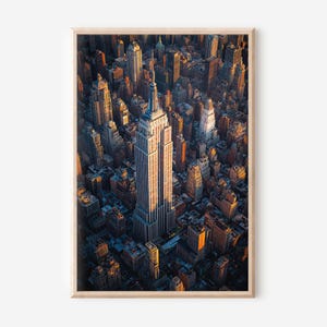 May include: An aerial view photograph of the New York City skyline, featuring the Empire State Building prominently. The image is framed in a light wood border, with the buildings bathed in warm sunlight, creating a contrast of light and shadow.