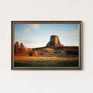 Könnte beinhalten: Gerahmter Landschaftsdruck mit Blick auf den Devils Tower in Wyoming. Das Bild zeigt den Turm unter blauem Himmel mit Wolken, umgeben von einer Wiese und Bäumen. Der Rahmen ist dunkelbraun.