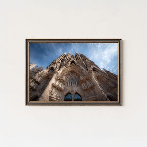 Op de afbeelding: Een ingelijste prent van de Sagrada Familia in Barcelona, Spanje. De afbeelding toont de architectonische details van de kathedraal tegen een blauwe lucht met wolken. De lijst is donker met een gouden binnenrand.