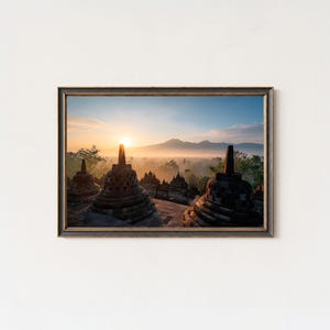 Könnte beinhalten: Gerahmter Druck eines Sonnenaufgangs über einer nebligen Landschaft mit alten Steinstupas. Die Sonne geht auf und wirft einen goldenen Schein über die Szene, mit Bergen in der Ferne. Das Kunstwerk befindet sich in einem dunkelbraunen Rahmen.