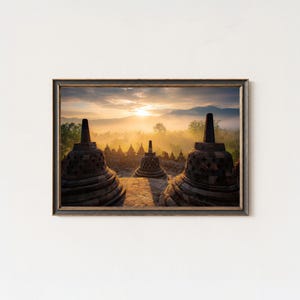 May include: Framed photograph of a temple complex at sunrise. The image features several bell-shaped stupas in the foreground, with a misty landscape and the sun in the background. The frame is dark brown.