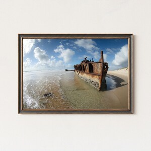 Op de afbeelding: Ingelijste print van een verroest scheepswrak op een zandstrand. De structuur van de boot is zichtbaar tegen een achtergrond van blauwe lucht en witte wolken. Het water spoelt zachtjes de kust.