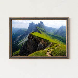 Puede incluir: Impresión enmarcada de un paisaje montañoso. La imagen muestra una cresta verde y herbosa con un sendero en primer plano, que conduce a una serie de montañas grises y azules bajo un cielo nublado. El marco es marrón oscuro.