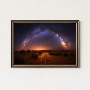 May include: Framed landscape print featuring a desert scene under a starry night sky. The Milky Way arches over the dunes, with a path of footprints leading towards the horizon. The frame is dark with a gold trim.