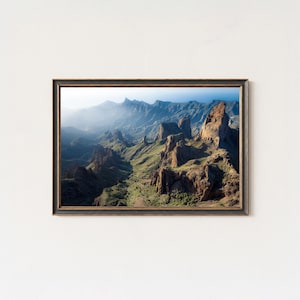 Op de afbeelding: Ingelijste landschapsafdruk met een bergketen met ruige toppen en weelderige groene valleien. Het kunstwerk toont een schilderachtig uitzicht met een blauwe lucht en zonlicht, waardoor een serene en natuurlijke esthetiek ontstaat. De lijst is donkerbruin.