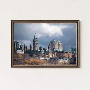May include: Framed print of a cityscape featuring a tall clock tower and various buildings under a cloudy sky. The buildings have green and brown roofs. The frame is dark with a gold trim.