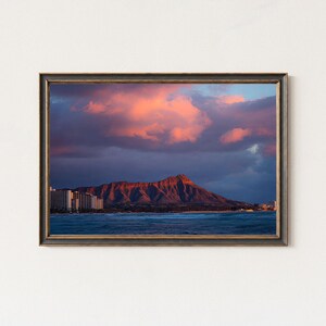 May include: Framed print of a Hawaiian landscape featuring Diamond Head. The image shows a sunset with pink and orange clouds above the mountain and ocean. Buildings are visible along the coast in the distance.