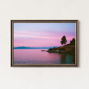May include: Framed landscape art print featuring a serene lake scene at sunset. The sky transitions from pink to purple, reflecting in the calm water. A small island with a tree and some bushes is on the right side of the image. The frame is dark brown.