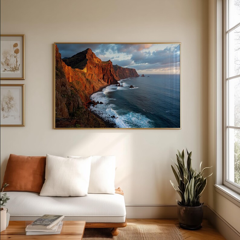 K&ouml;nnte beinhalten: Gerahmte Landschaftsfotografie einer K&uuml;stenlandschaft. Das Bild zeigt eine zerkl&uuml;ftete, rotbraune Felswand, die auf den tiefblauen Ozean trifft. Der Himmel ist eine Mischung aus Blau und Grau, mit einem Hauch von Orange von der untergehenden Sonne. Das Foto ist in einem goldenen Rahmen.