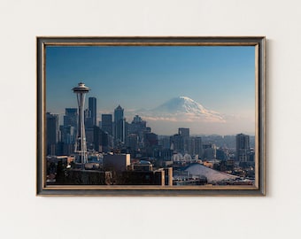 May include: Framed print depicting the Seattle skyline, with the Space Needle and a snow-covered mountain visible. The city's buildings are set against a clear blue sky. The frame has a dark, ornate design.