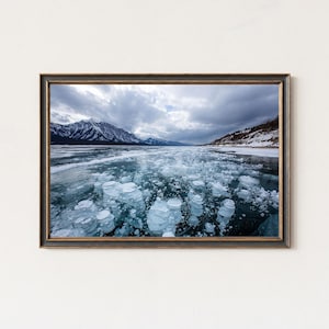 May include: A framed photograph of a frozen lake with ice bubbles. The image features a landscape with mountains in the background and a cloudy sky. The frame is dark brown.