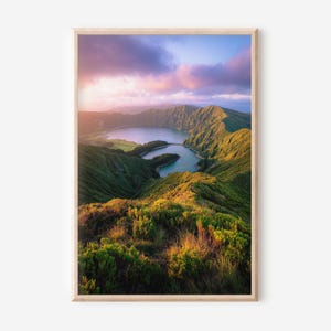May include: A framed landscape print showing a lake nestled in green hills under a sky of pink and purple clouds. The sunlight bathes the scene in a warm light. The print is presented in a natural wood frame.