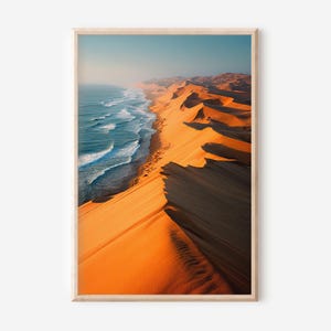 May include: A framed print showcasing a coastal landscape. The image features a vibrant contrast between the blue ocean waves and the orange sand dunes. The sky is a soft blue, and the scene is bathed in sunlight, creating a warm and inviting atmosphere.