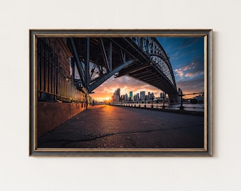 May include: Framed photograph of the Sydney Harbour Bridge at sunset. The bridge's steel structure frames the cityscape and the vibrant colors of the sky. The image captures the golden hour, with the sun's glow reflecting on the water and pavement.