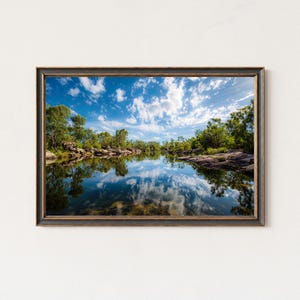 Puede incluir: Una impresión enmarcada de un paisaje fluvial sereno. La imagen captura un cielo azul vibrante salpicado de nubes blancas y esponjosas reflejadas en el agua tranquila. Frondosos árboles verdes bordean las orillas.