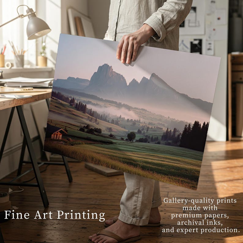 A person holding a large framed print of a scenic landscape with mountains and a small village in the foreground.