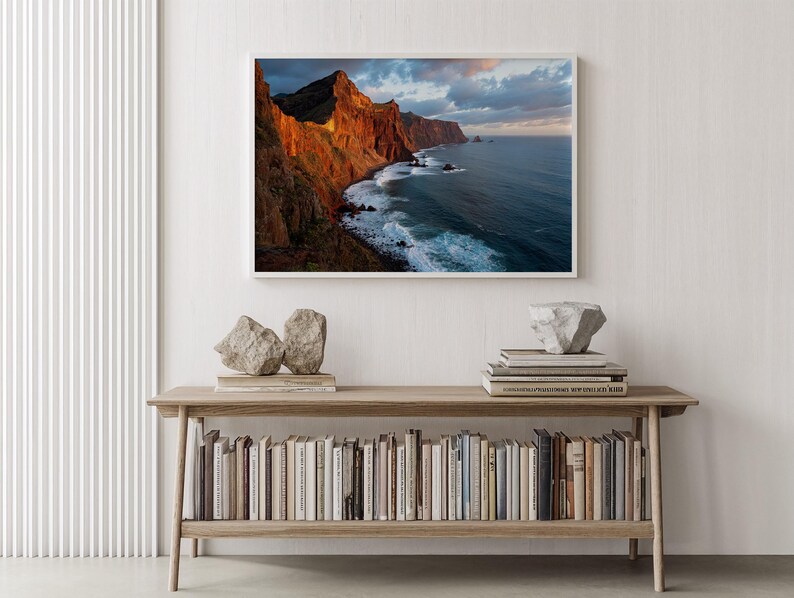 K&ouml;nnte beinhalten: Gerahmte Landschaftsfotografie einer K&uuml;stenlandschaft mit einem Gebirgszug. Die Berge sind rotbraun und das Meer tiefblau. Das Foto befindet sich &uuml;ber einer Holzkonsolentisch mit B&uuml;chern und dekorativen Steinen.
