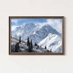May include: Framed landscape art print featuring a snow-covered mountain range under a blue sky with scattered clouds. The artwork includes dark green trees on the mountainside and is encased in a dark brown frame.