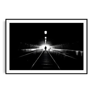Op de afbeelding: Zwart-wit ingelijste print van een persoon die een fel verlichte tunnel inloopt. De tunnel heeft lichten langs het plafond en een rolpad. De afbeelding speelt met contrasten, met de donkere tunnel die naar een helder licht leidt.