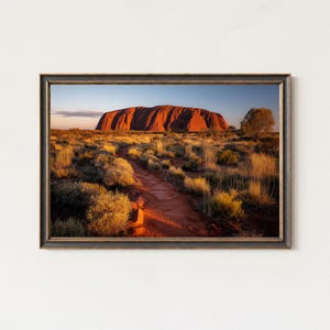 May include: Framed print of a desert landscape with a large, red rock formation. A dirt path winds through the foreground, flanked by dry vegetation and small trees. The sky transitions from blue to orange.