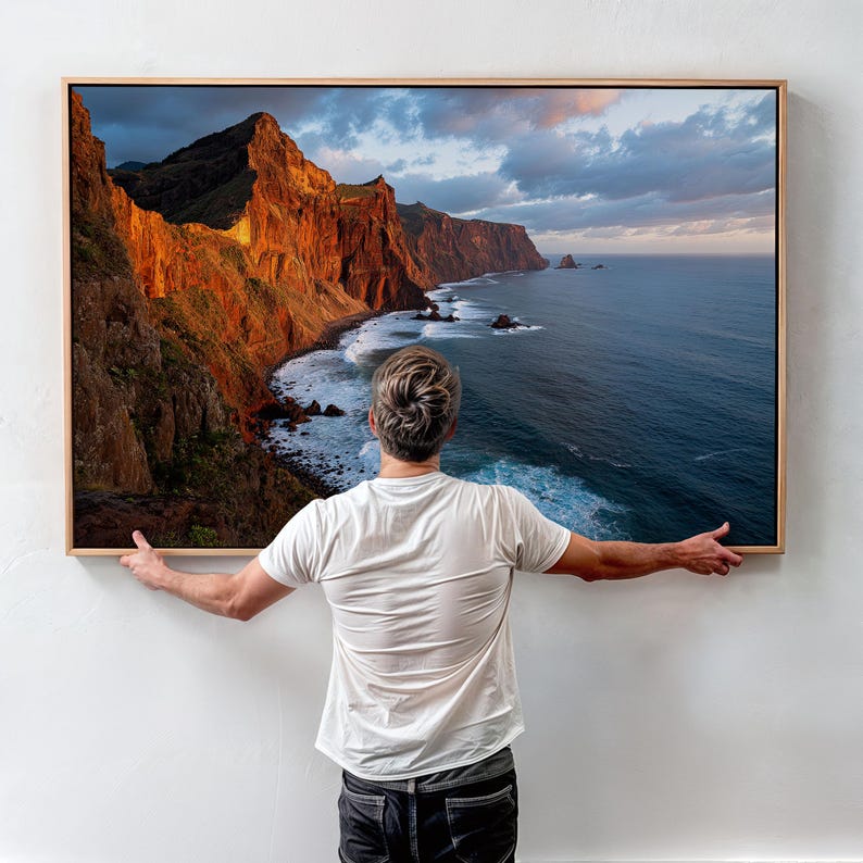 K&ouml;nnte beinhalten: Ein gerahmter Landschaftsdruck mit einer K&uuml;stenlandschaft mit Klippen und Meer. Das Kunstwerk wird von einer Person gehalten, die ein wei&szlig;es T-Shirt und dunkle Jeans tr&auml;gt. Die Klippen werden von der Sonne beleuchtet, das Meer in Blaut&ouml;nen.