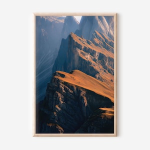 May include: A framed print featuring a mountain landscape. The image showcases rugged, layered mountain peaks in shades of brown and blue, illuminated by sunlight. The artwork is encased in a light-colored wooden frame, suitable for home decor.