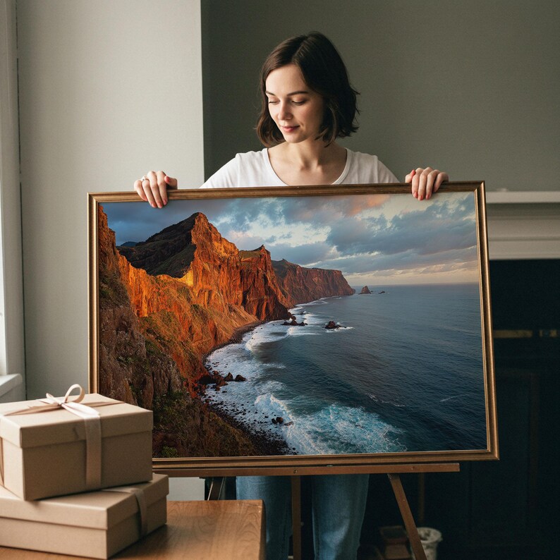 K&ouml;nnte beinhalten: Ein gerahmter Landschaftsdruck mit einer K&uuml;stenlandschaft mit dramatischen Klippen und Meereswellen. Das Kunstwerk hat einen goldfarbenen Rahmen. Die Klippen werden von warmem Sonnenlicht beleuchtet, das mit den k&uuml;hlen T&ouml;nen des Meeres und des Himmels kontrastiert. Zwei braune Geschenkboxen stehen auf einem Holztisch.
