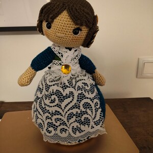 May include: A handmade crochet doll with a tan face and brown hair styled in a bun. The doll wears a blue and white dress with a white lace overlay and a gold-colored jewel.