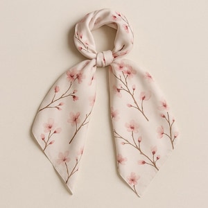 Könnte beinhalten: Ein weißer Seidenschal mit einem zarten rosafarbenen Blumendruck von Kirschblüten. Der Schal ist zu einem Knoten gebunden und hat eine dreieckige Form.