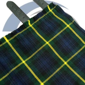 May include: Close-up of a traditional tartan kilt with a green, navy blue, and yellow plaid pattern. The kilt has a fringed edge and two dark gray leather straps.