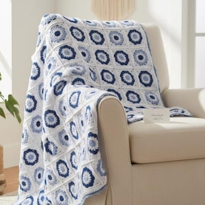 May include: A crochet blanket featuring a repeating pattern of circular floral motifs in shades of navy blue, light blue, and white. The squares are joined with white yarn, creating a grid structure. A small white card reads Handmade with love.