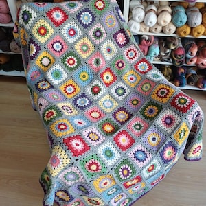 May include: A colorful, handmade crochet blanket draped over a chair. The blanket features a patchwork design of square motifs in various colors, including red, yellow, green, and blue, with a gray border. The blanket is made of yarn.