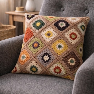 May include: A square crochet cushion with a geometric pattern of colourful squares. The squares feature floral designs in shades of orange, green, yellow, and brown. The cushion is resting on a grey armchair.