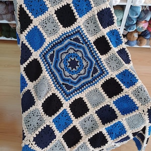 Puede incluir: Una manta de ganchillo hecha a mano en tonos azules, negros, grises y crema. La manta presenta un diseño central en forma de estrella rodeado de motivos cuadrados. Los cuadrados están dispuestos en un patrón de cuadrícula, creando un diseño geométrico.