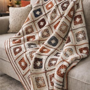 Può includere: Una coperta all'uncinetto fatta a mano, drappeggiata su un divano. La coperta presenta un motivo quadrato con quadrati alternati color crema, marrone e blu. I bordi della coperta sono rifiniti con un bordo marrone, creando un'estetica accogliente.