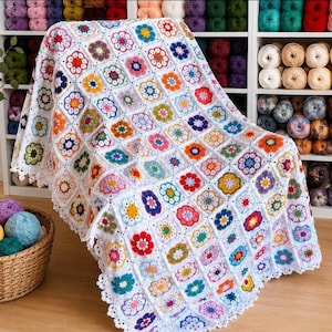 May include: A handmade, white crochet blanket with a colorful floral pattern. The blanket features square motifs with flowers in various colors, including red, blue, yellow, and pink. The blanket is draped over a chair and has a scalloped edge.