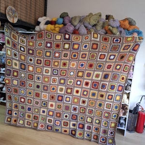 May include: A large, handmade crochet blanket with a patchwork design. The blanket features a brown border and is filled with colorful squares in shades of red, yellow, blue, and green. The blanket is draped over a shelf filled with yarn.
