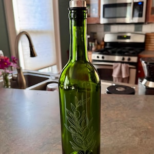 May include: A green glass olive oil bottle with a stainless steel spout. The bottle has a decorative etched olive branch design. The bottle is on a countertop in a kitchen setting.