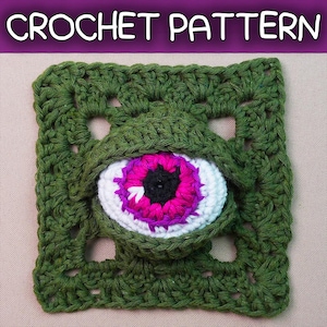 May include: A square crochet pattern featuring a dark olive green granny square base with a three-dimensional eye motif in the center. The eye has a black pupil, a vibrant magenta iris, and a white sclera, all framed by green crochet stitches.