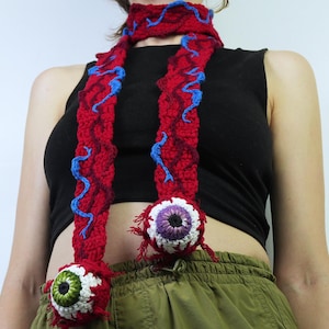 K&ouml;nnte beinhalten: Roter H&auml;kelschal mit blauen Akzenten und zwei gro&szlig;en, detaillierten augenf&ouml;rmigen Ornamenten. Der Schal wird um den Hals getragen. Die Augen haben wei&szlig;e, violette und gr&uuml;ne Details. Einzigartiges Accessoire.