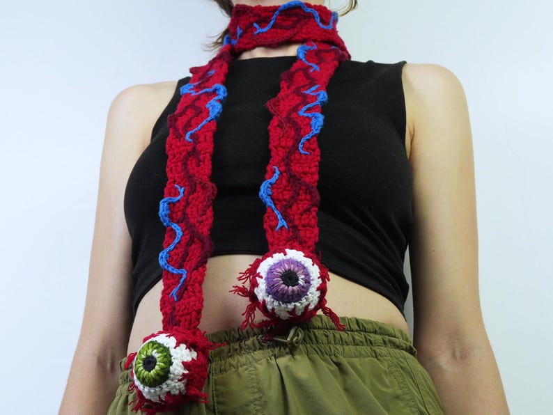K&ouml;nnte beinhalten: Roter H&auml;kelschal mit blauen Akzenten und zwei gro&szlig;en, detaillierten augenf&ouml;rmigen Ornamenten. Der Schal wird um den Hals getragen. Die Augen haben wei&szlig;e, violette und gr&uuml;ne Details. Einzigartiges Accessoire.